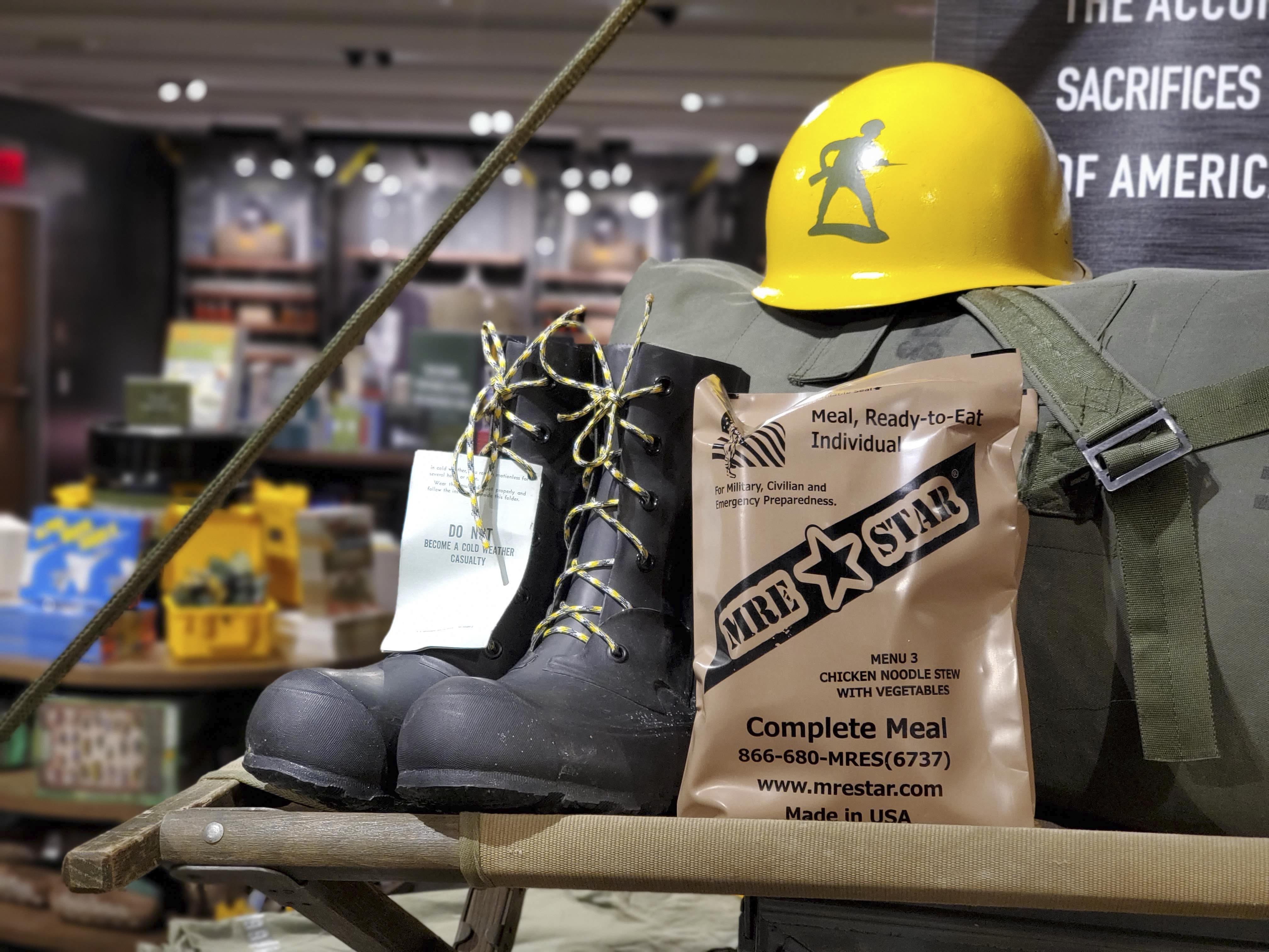 A field-kit vignette — cold-weather boots, MRE, and helmet staged on a folding cot